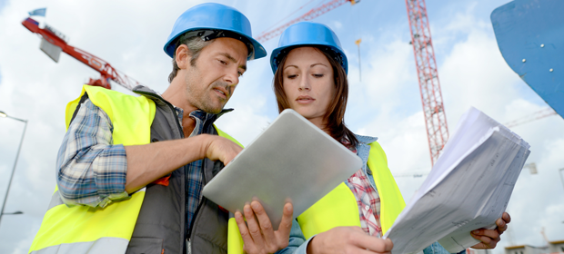 Dieses Bild zeigt einen Architekten und eine Architektin mit Bauhelm und Warnwesten, die sich mit Tablet PC und Bauplan besprechen (© goodluz - Fotolia.com / 44575141)
